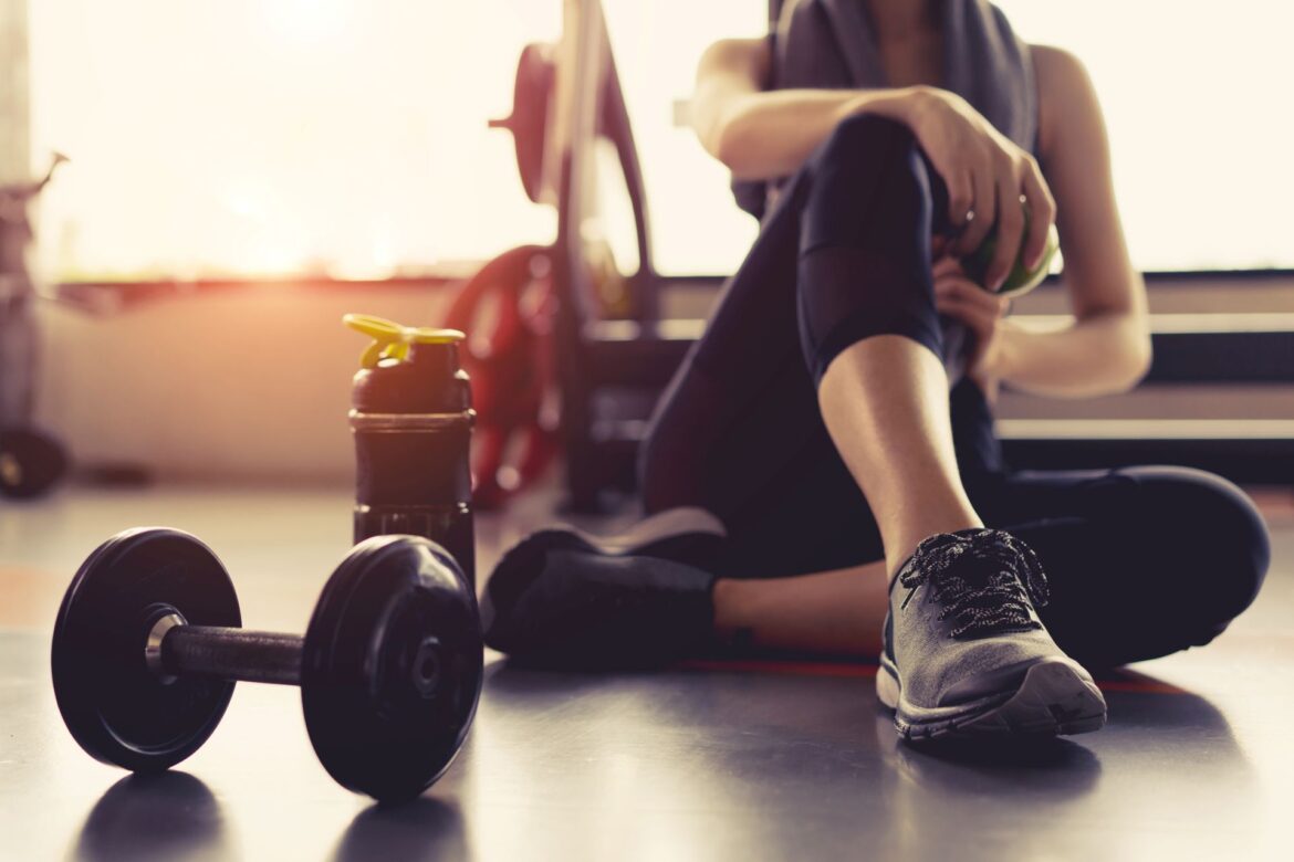 Woman relaxing after a gym workout with dumbbells, an apple, and a protein shake—using exercise for public speaking anxiety and stress recovery.