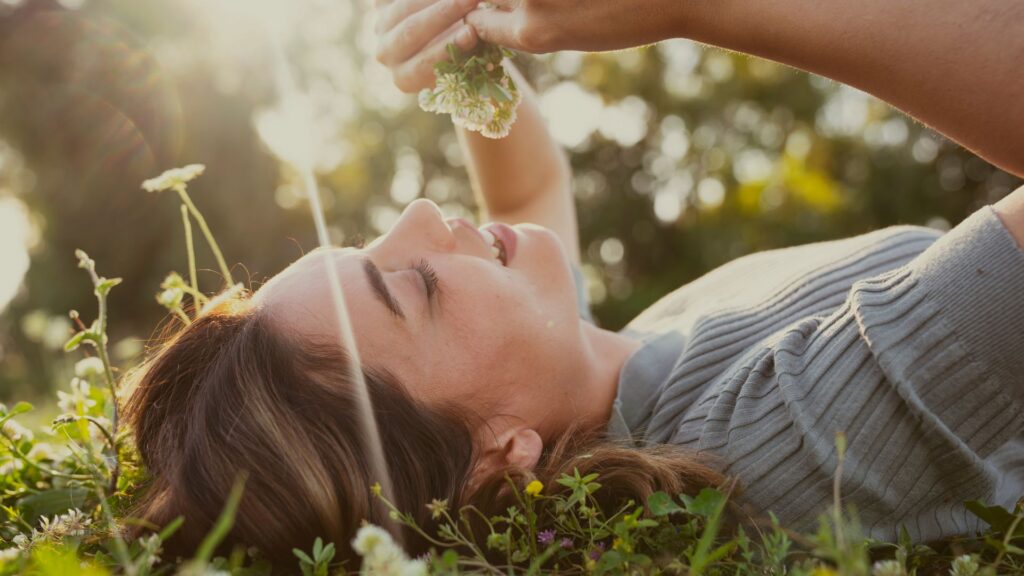 A woman lying in a field, she smells a flower, fully present in the moment. A peaceful reminder of how to be more present daily.