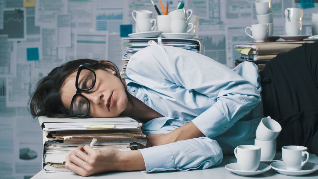 Why you need to pause – a woman asleep at her desk, surrounded by coffee cups and paperwork, illustrating exhaustion, overwork, and the need for intentional breaks.