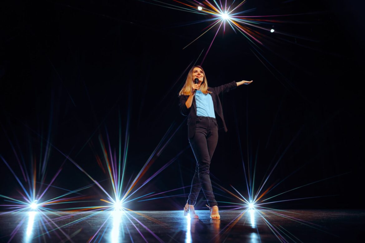 A confident woman standing on stage, commanding attention through a strategic pause, demonstrating how to use pauses in public speaking to engage her audience.